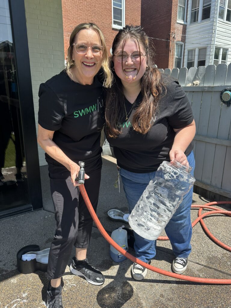Two women smiling outdoors, both wearing black t-shirts and glasses. One woman is holding a red hose nozzle, and the other is holding a large clear plastic container filled with soapy water. They are standing on a concrete surface near a gray wooden fence and brick buildings. Various cleaning supplies are visible on the ground nearby.