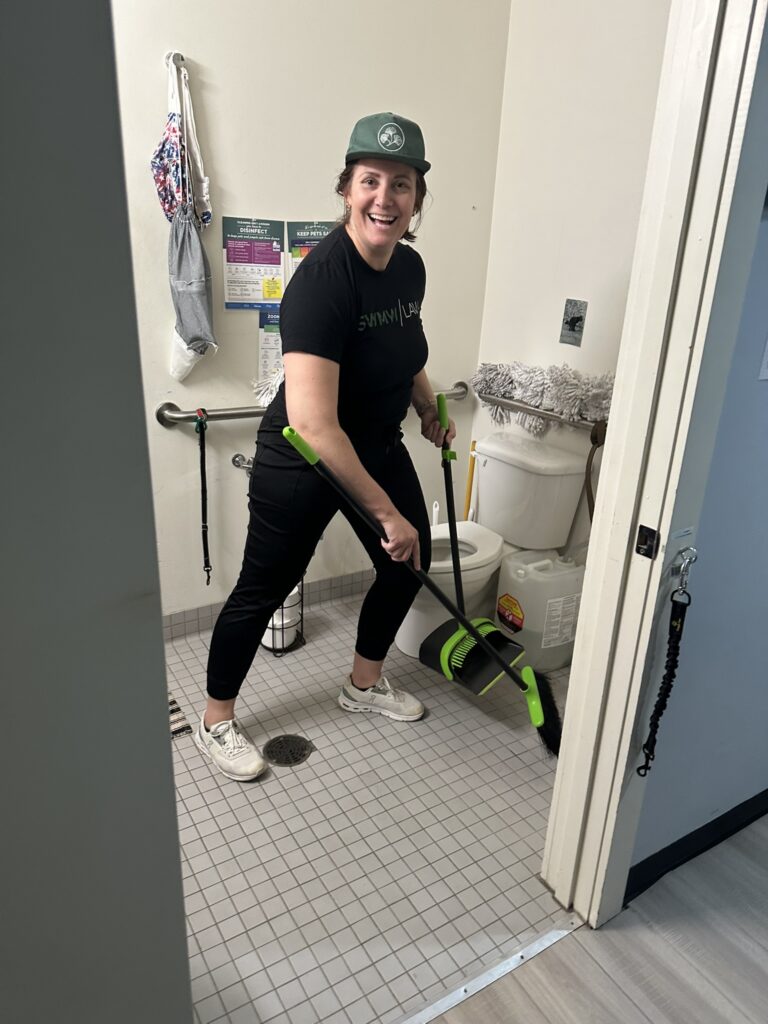 A woman wearing a green cap, black t-shirt, black pants, and white sneakers is sweeping the floor of a small tiled bathroom with a green and black broom and dustpan. The bathroom has a white toilet, a mop hanging on a grab bar, and various cleaning supplies. The woman is smiling and looking toward the camera.