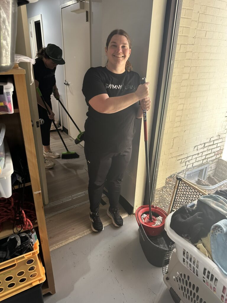 A person in black clothing is standing indoors holding a mop with a red mop bucket nearby. Another person in black clothing and a dark green cap is sweeping the floor in the background. The room has a light-colored floor and walls, with a laundry basket filled with clothes and a shelving unit with various items on the left side.