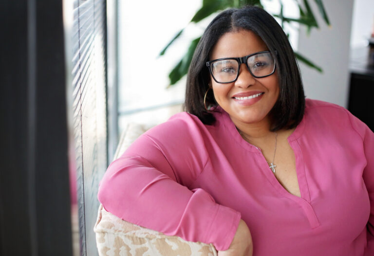 A woman with shoulder-length black hair wearing large black-rimmed glasses and a pink blouse is smiling while leaning on a beige patterned cushion. She is also wearing a silver necklace with a cross pendant and gold hoop earrings. The background includes a window with blinds and some green plant leaves.