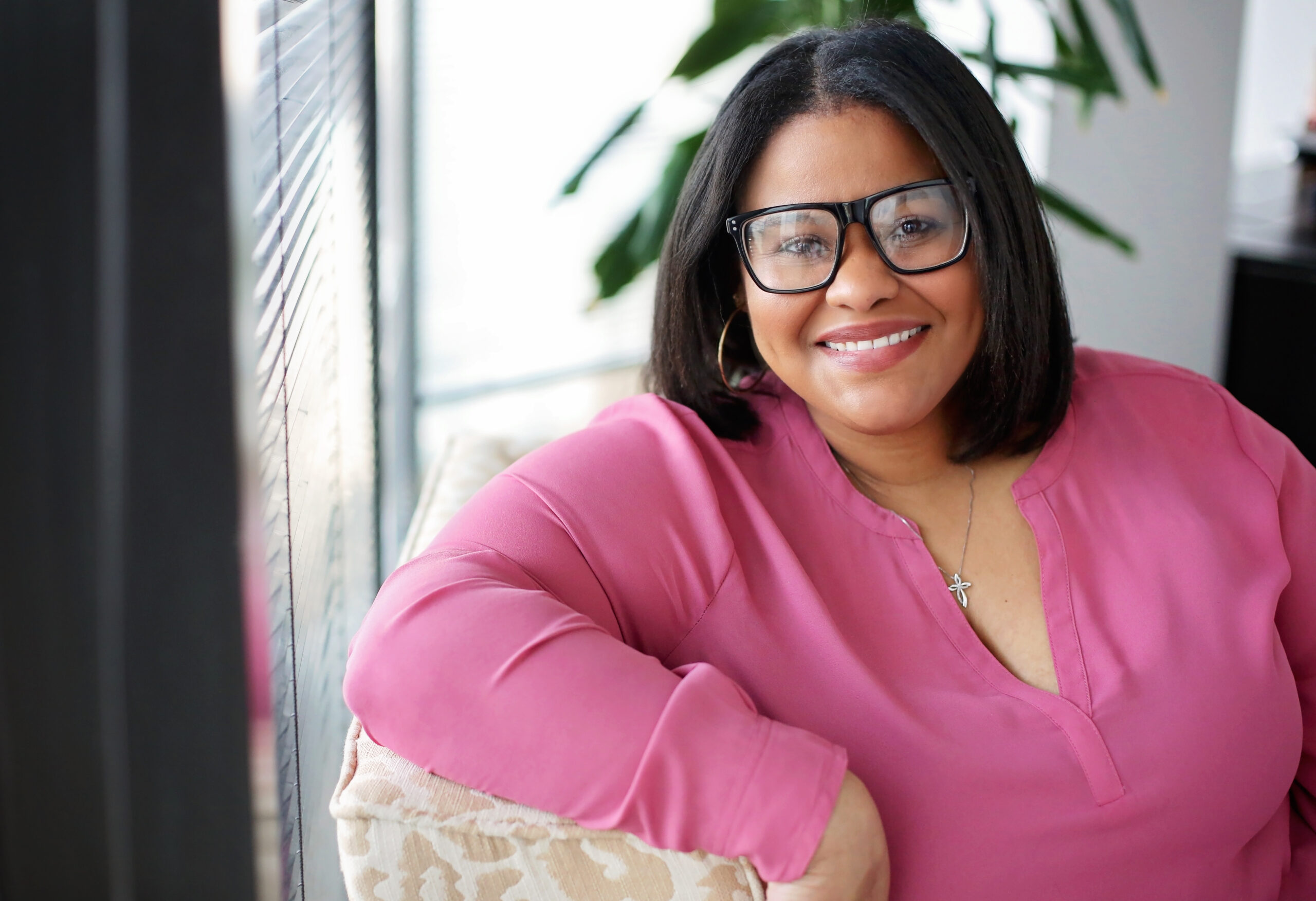 A woman with shoulder-length black hair wearing large black-rimmed glasses and a pink blouse is smiling while leaning on a beige patterned cushion. She is also wearing a silver necklace with a cross pendant and gold hoop earrings. The background includes a window with blinds and some green plant leaves.