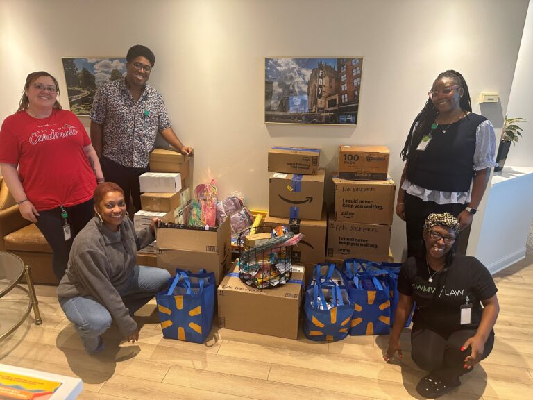 Five people are posing indoors around a collection of stacked cardboard boxes, colorful backpacks, and blue reusable bags filled with supplies. The group is smiling, with two individuals standing, two kneeling, and one crouching. The setting includes a wooden floor, a brown leather couch, and framed pictures on the wall. The boxes are labeled with phrases like "Kids Backpacks" and "100 Pack Colored Pencils.